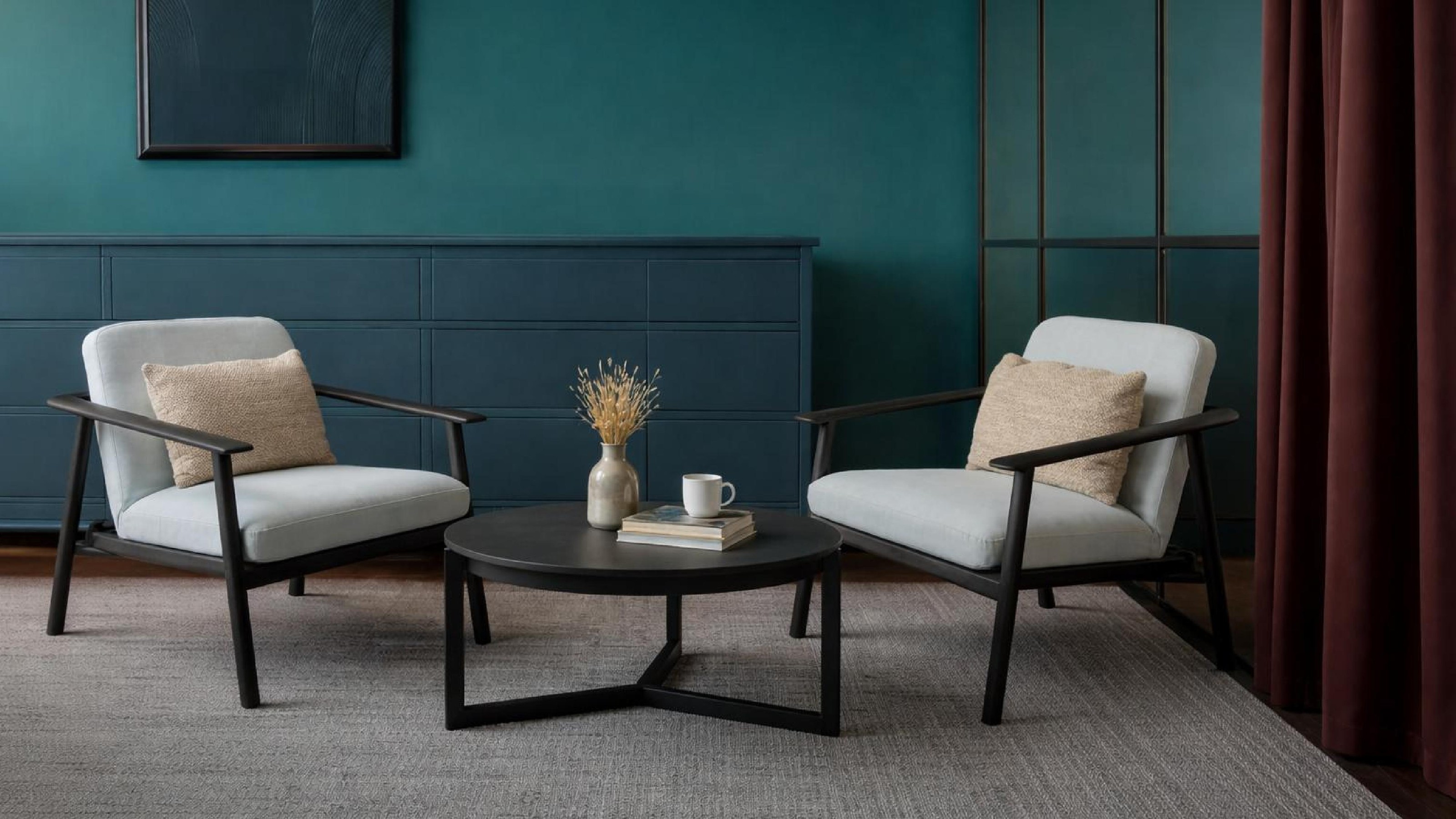 Two grey lounge armchairs and a round table against a deep teal panelled wall